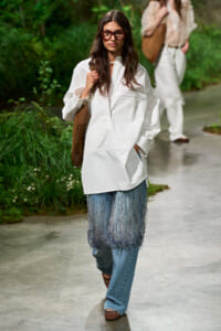 Model wearing an oversized white shirt with a fringe denim layer over blue jeans, brown tote, and red-framed sunglasses walking down an outdoor runway with greenery in the background.