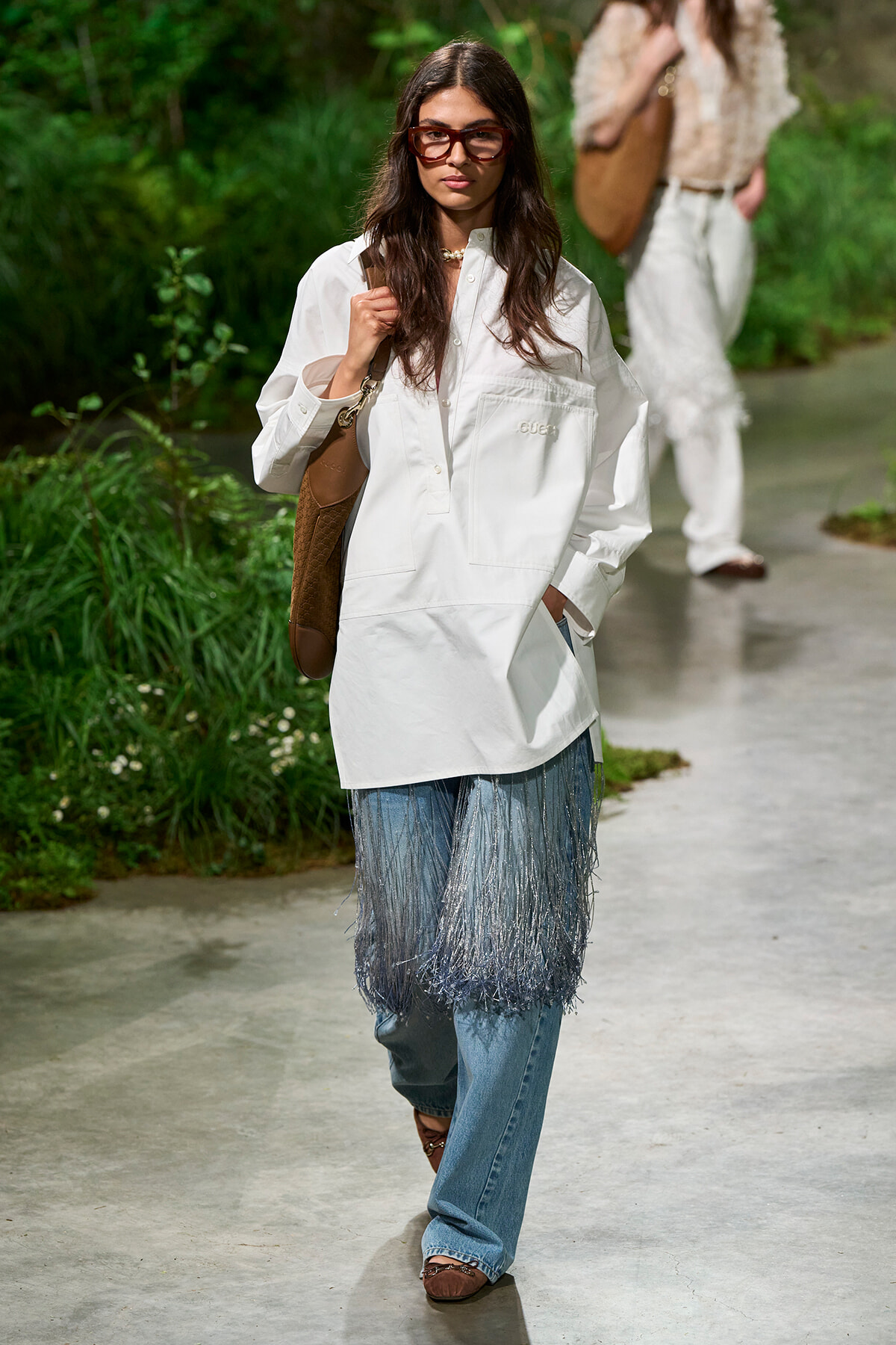 Model wearing an oversized white shirt with a fringe denim layer over blue jeans, brown tote, and red-framed sunglasses walking down an outdoor runway with greenery in the background.