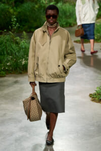 Model walking a garden runway in a tan zip-front jacket, dark midi skirt, sunglasses, pearl choker, and carrying a beige patterned handbag.