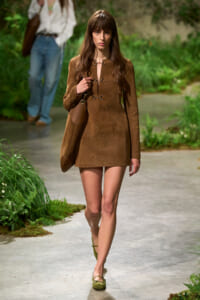 Female model walking an outdoor runway in a brown textured mini dress with a matching handbag and green flats, surrounded by greenery on the catwalk.