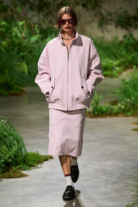 Model in pale pink oversized jacket and midi skirt walking on a garden runway, wearing dark sunglasses and a pearl necklace.