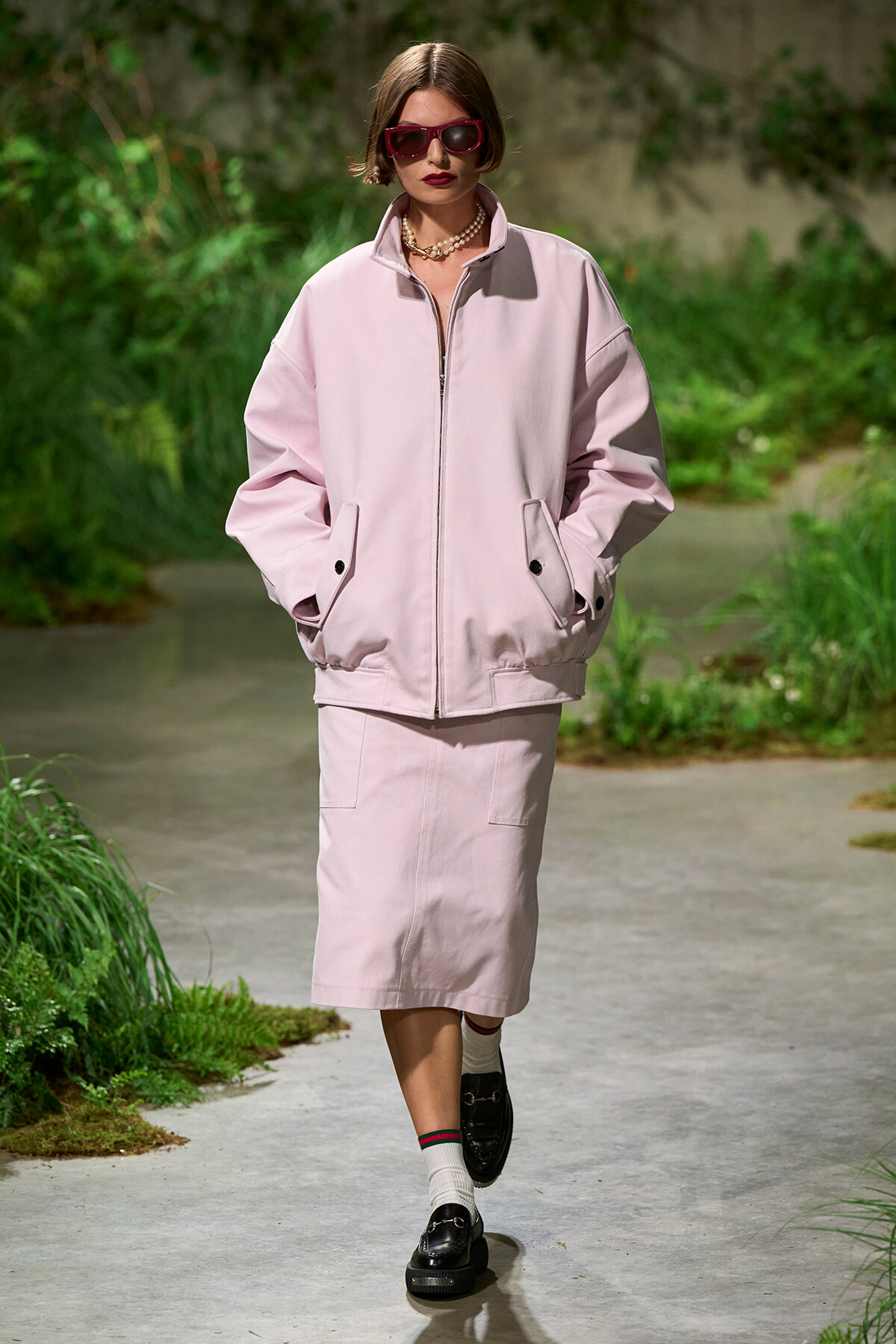 Model in pale pink oversized jacket and midi skirt walking on a garden runway, wearing dark sunglasses and a pearl necklace.