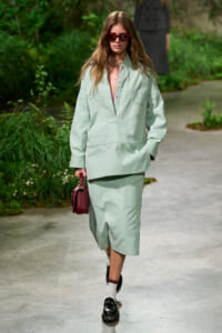 Model in a mint-green oversized shirt and skirt with a burgundy handbag and red sunglasses walking on an outdoor runway court.