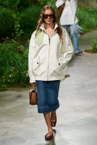 Model walks a runway in a cream zip-front jacket with oversized collar, a blue denim midi skirt, red sandals, and a brown handbag.