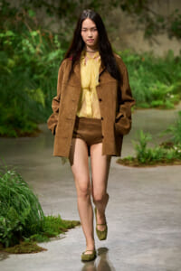 Model walking along a garden path in a brown plaid coat, yellow blouse, brown shorts, and green flats.