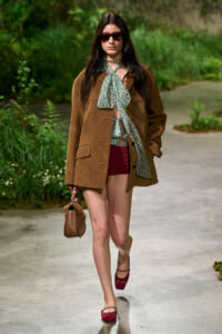 Woman walking outdoors in a brown textured blazer, teal floral scarf, burgundy shorts, red flats, sunglasses, carrying a tan handbag.