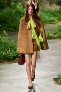Model walking an outdoor garden runway in a camel cape over a lime green blouse, brown shorts, and a burgundy handbag.