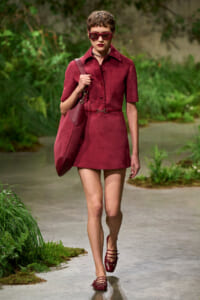 Model walking along a garden path in a burgundy shirt dress with a matching burgundy bag and sunglasses.