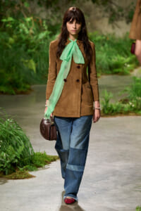 Model walks a runway in a brown suede coat with a mint-green scarf, wide-leg denim jeans, and a brown handbag.