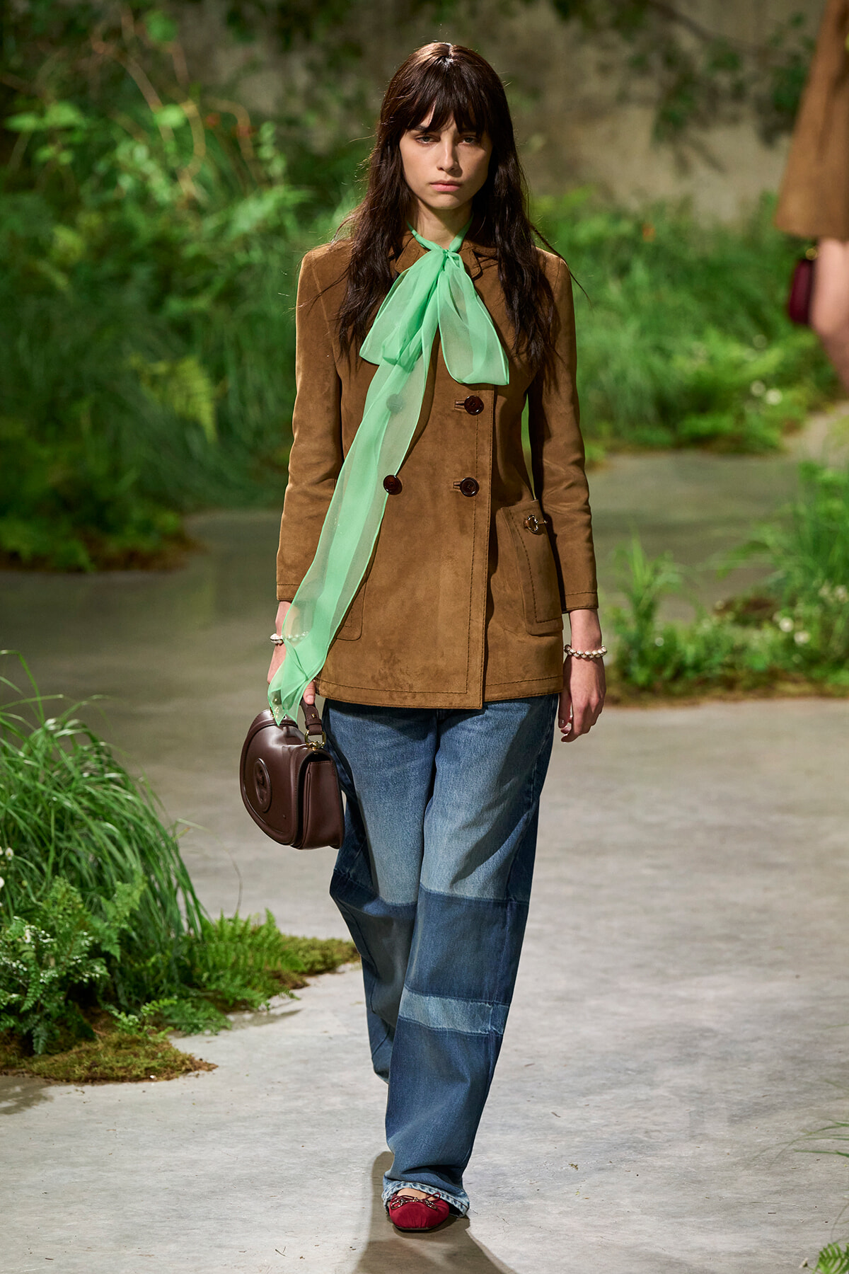 Model walks a runway in a brown suede coat with a mint-green scarf, wide-leg denim jeans, and a brown handbag.