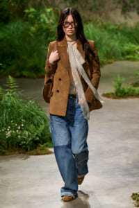 Woman walking outdoors in a brown textured blazer, light patterned scarf, blue denim jeans, and brown shoes with a brown handbag. She wears sunglasses and a necklace.