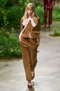 Model walking a runway in a brown suede-like suit with a pale pink scarf tied in a bow at the neck, outdoors among greenery.