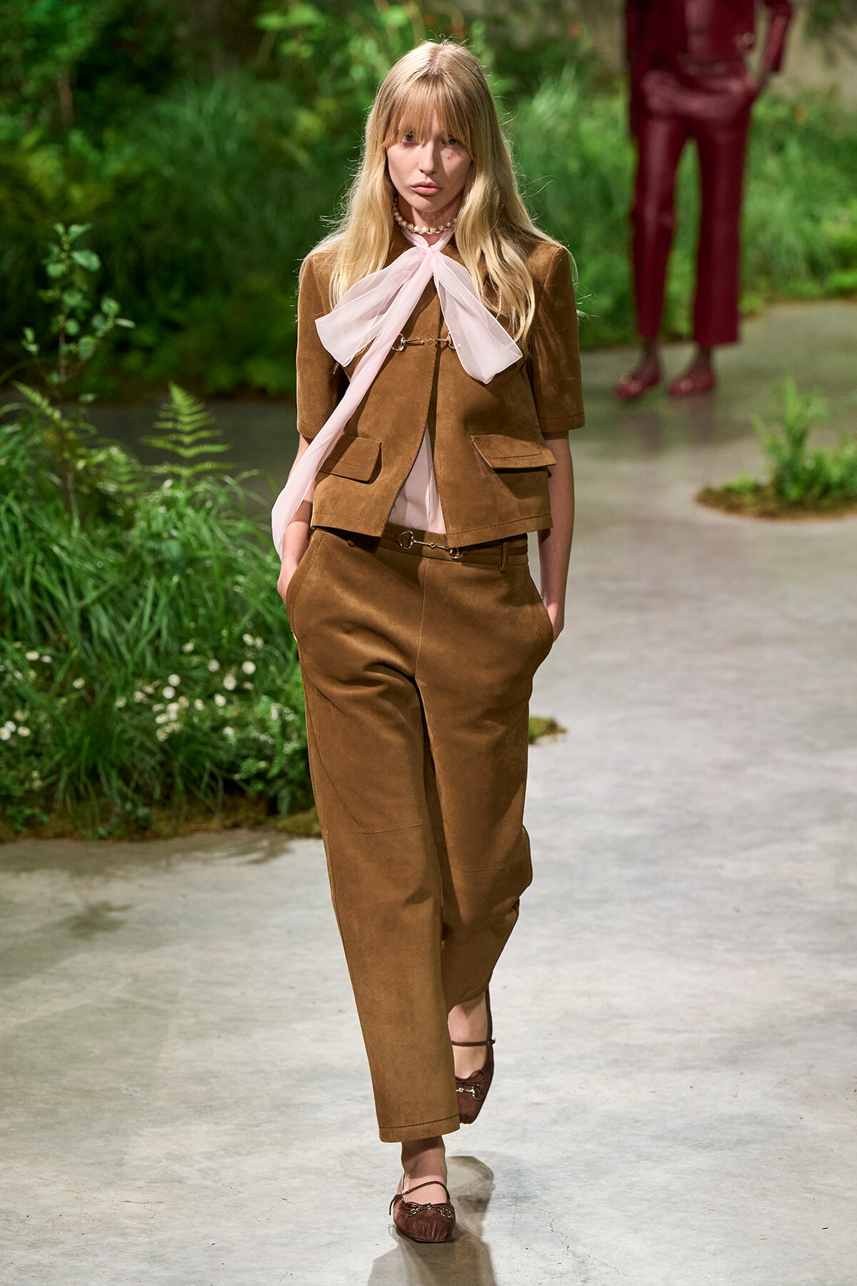 Model walking a runway in a brown suede-like suit with a pale pink scarf tied in a bow at the neck, outdoors among greenery.
