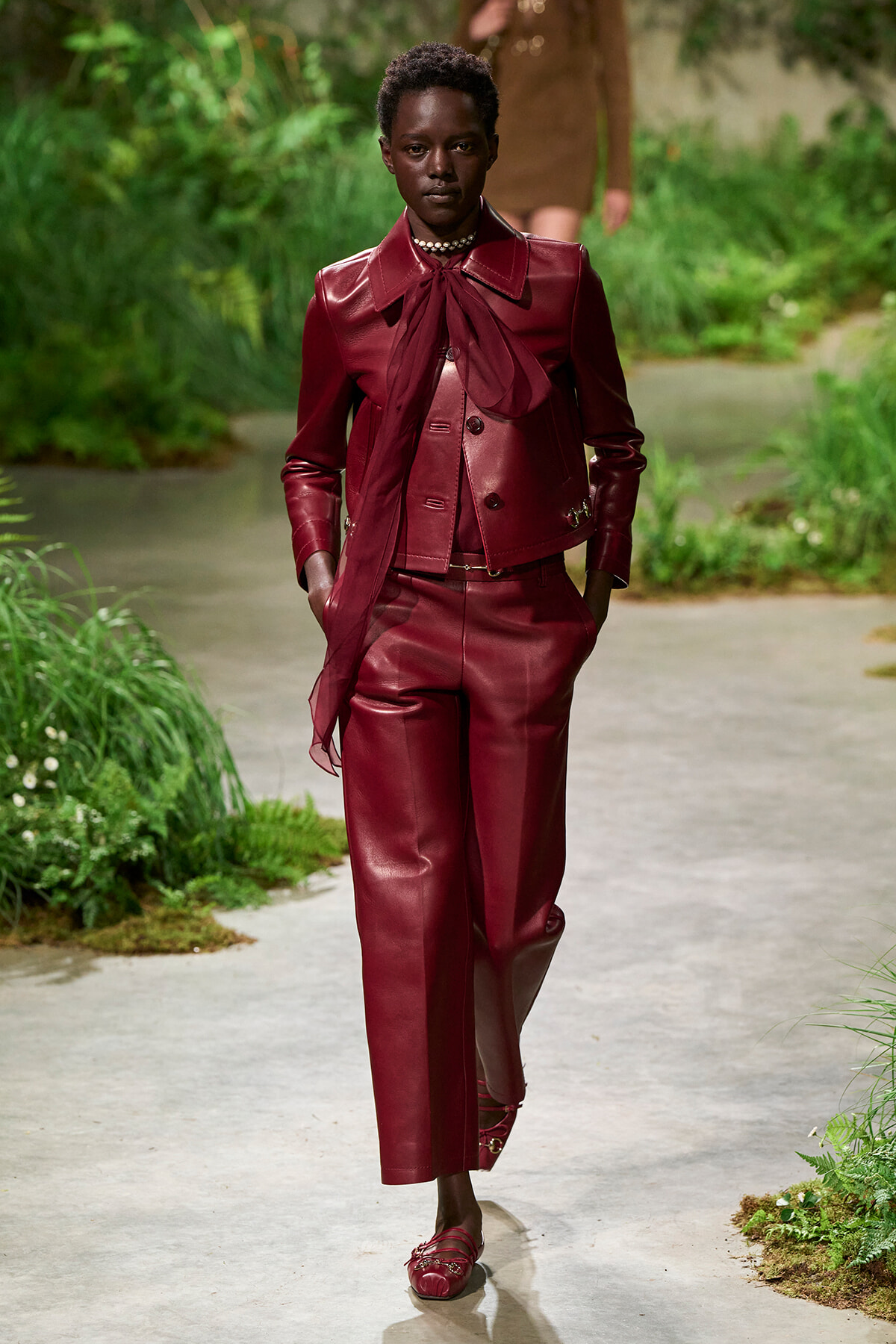 Model walks down a garden runway wearing a burgundy leather suit with a attached scarf bow and matching loafers.