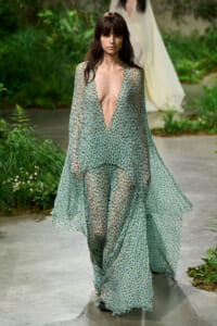 Model walking an outdoor runway in a flowing green floral sheer gown with a deep V-neck.