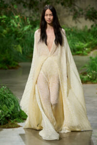 Model walking a runway in a cream, sheer dotted gown with a deep V-neck and flowing cape-like overlay.