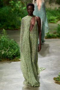 Model walking a runway in a floor-length green floral dress with a deep side cutout, outdoors in a garden setting.