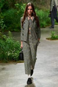 Model walking a garden-themed runway in a matching black-and-white floral suit, carrying a small black handbag.