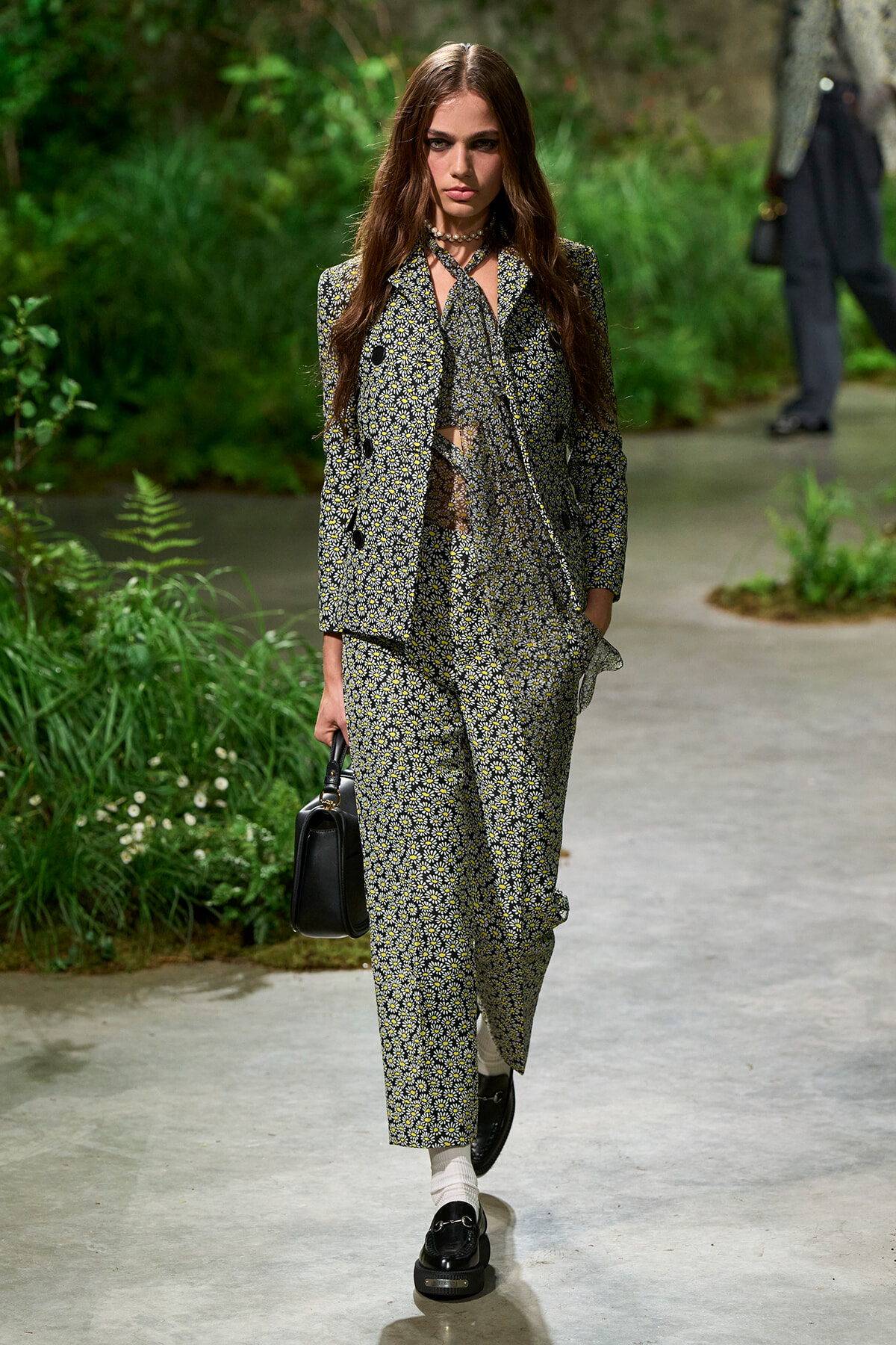 Model walking a garden-themed runway in a matching black-and-white floral suit, carrying a small black handbag.