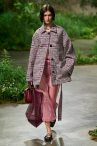 Female model walking on a garden path in a pink plaid oversized coat, sheer pink pants, black flats, and a burgundy handbag with a mesh tote.