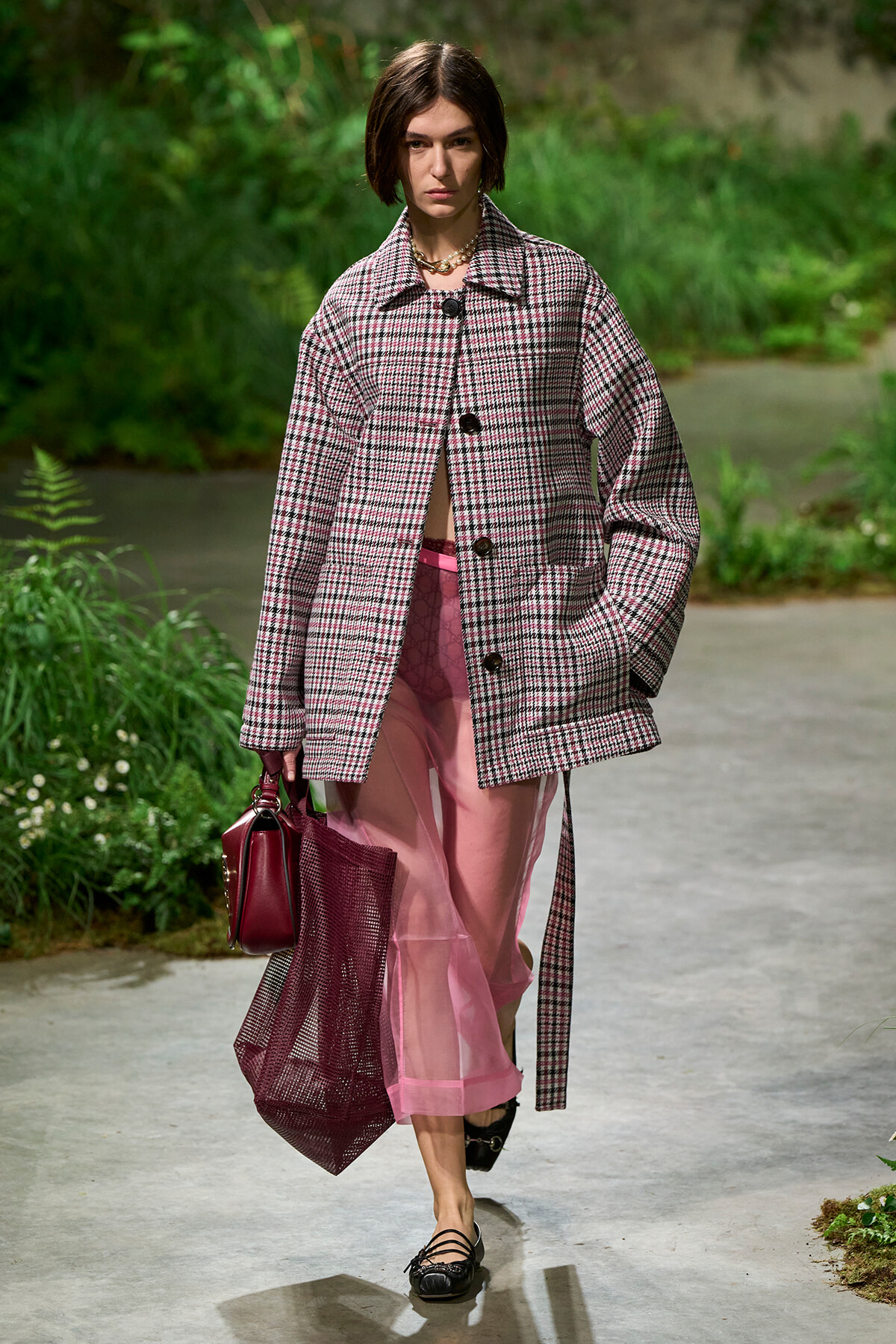 Female model walking on a garden path in a pink plaid oversized coat, sheer pink pants, black flats, and a burgundy handbag with a mesh tote.