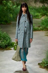 Woman walking along a garden path wearing a blue plaid coat, teal sheer pants, and red flats, carrying a green handbag and beige mesh tote.