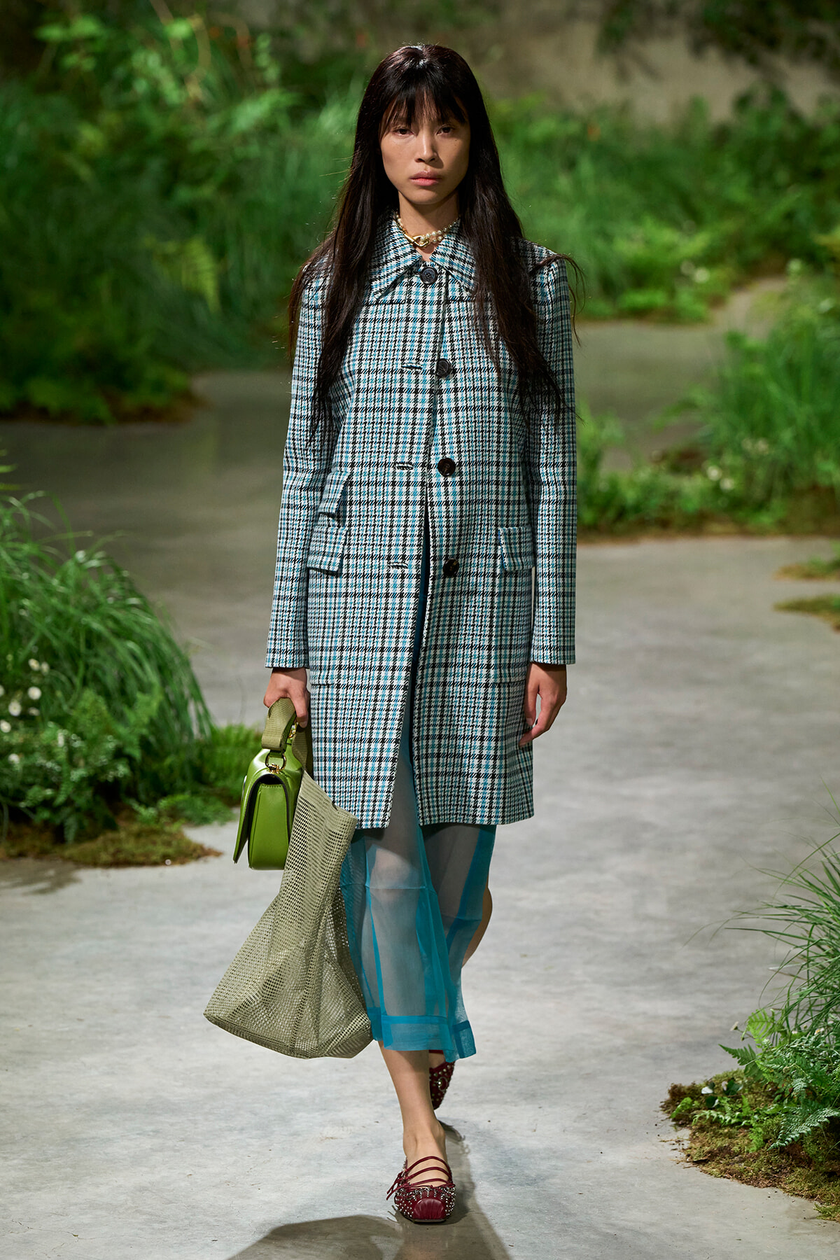 Woman walking along a garden path wearing a blue plaid coat, teal sheer pants, and red flats, carrying a green handbag and beige mesh tote.