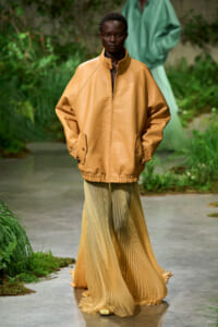 Model wearing oversized tan jacket with high collar and elastic hem, paired with flowing pleated champagne skirt on an outdoor garden runway