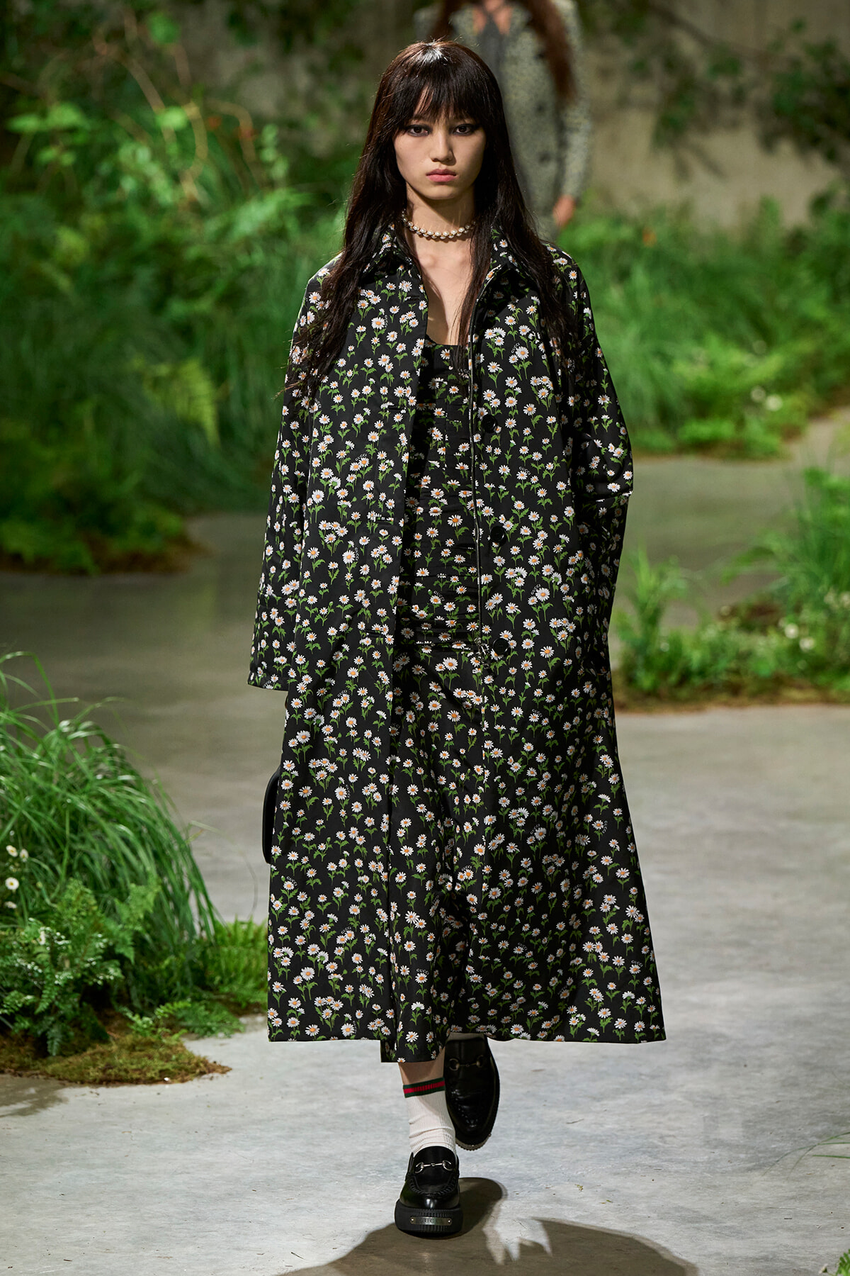 Female model walking an outdoor runway, wearing a black daisy-print coat over a matching dress with white flowers.