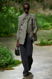 Model wearing a daisies-pattern blazer, dark jeans, sunglasses, pearl necklace, and holding a small black handbag on a garden path.