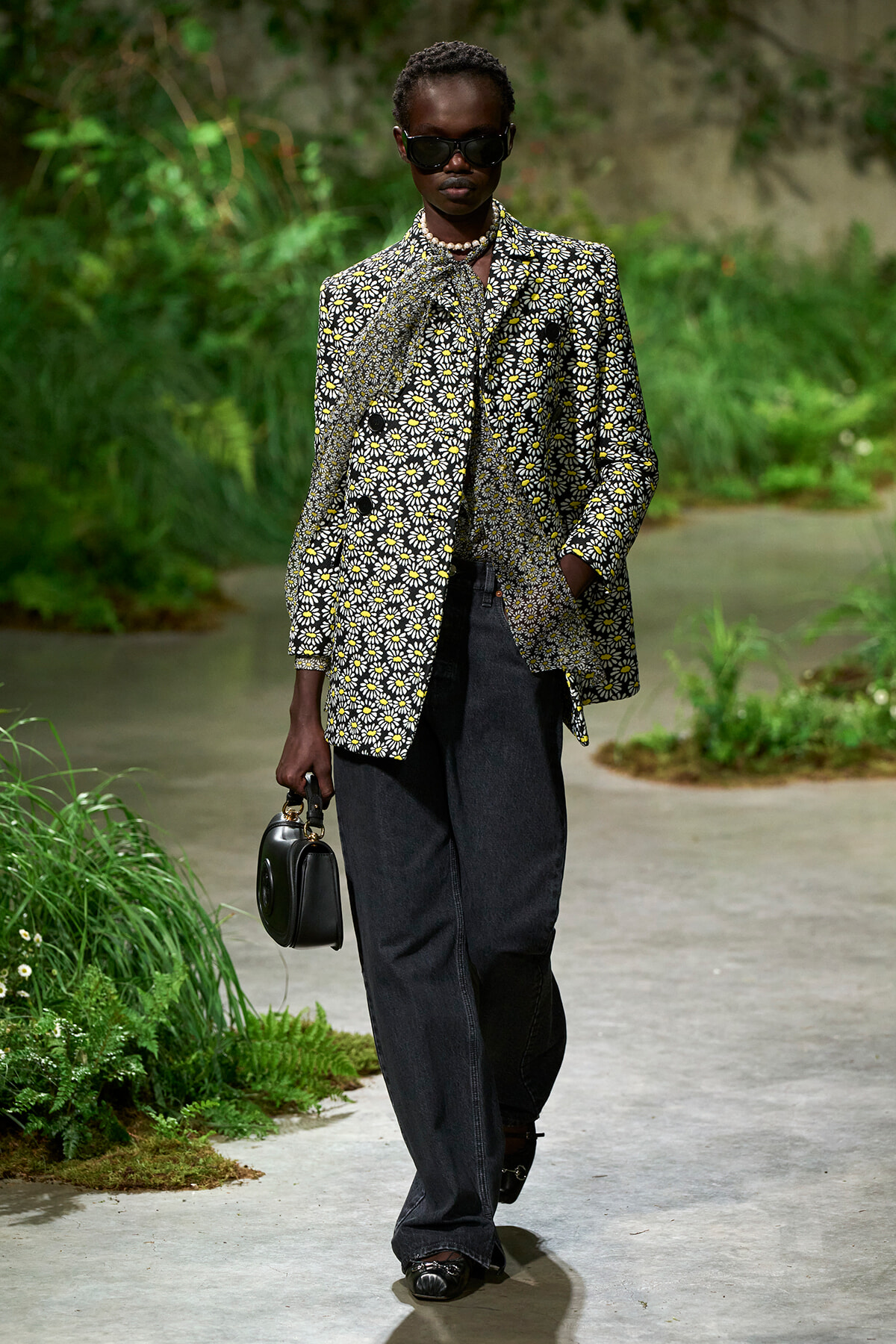 Model wearing a daisies-pattern blazer, dark jeans, sunglasses, pearl necklace, and holding a small black handbag on a garden path.