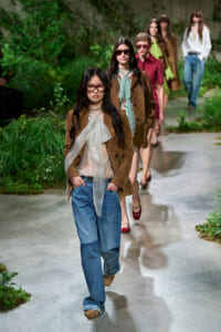 Model wearing a brown suede blazer, light blouse with a neck scarf, and wide-leg jeans walks down an outdoor garden runway.