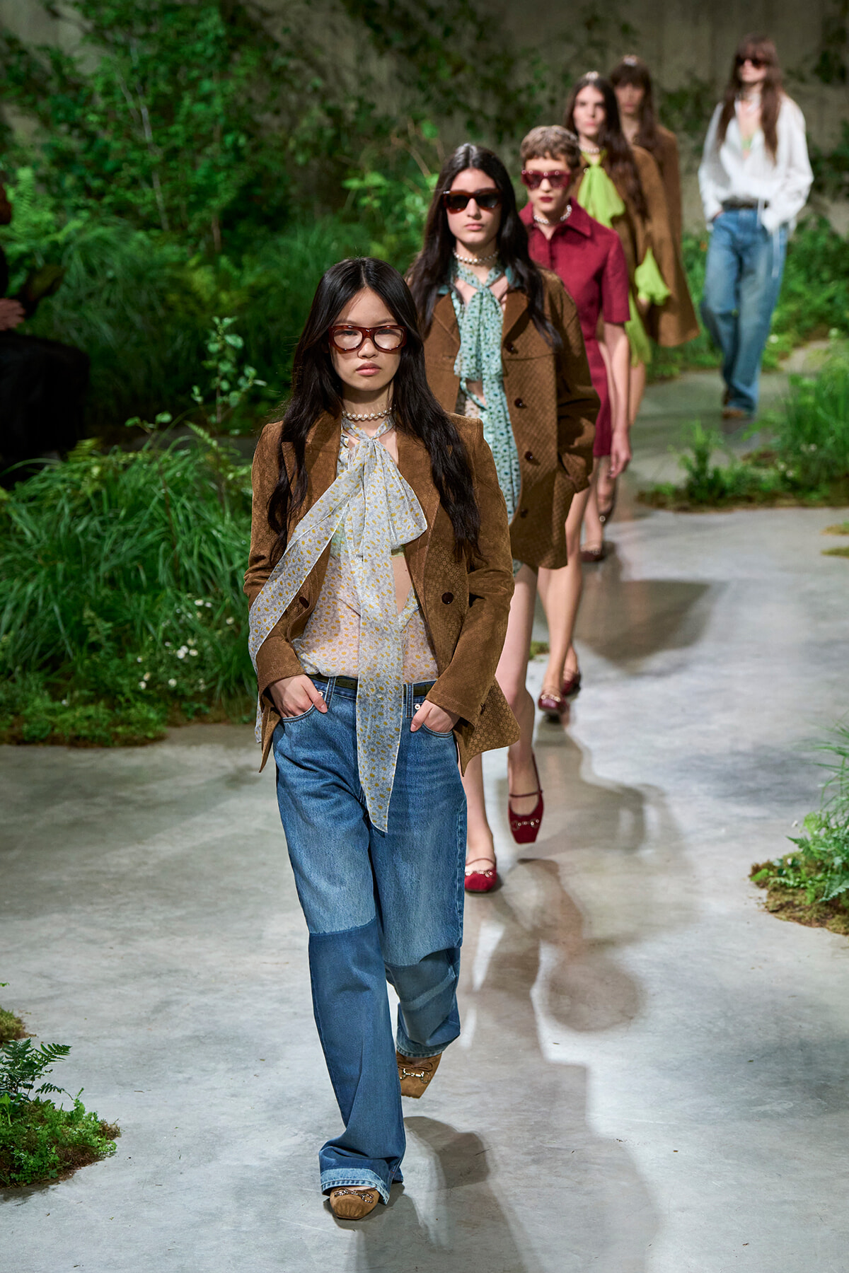 Model wearing a brown suede blazer, light blouse with a neck scarf, and wide-leg jeans walks down an outdoor garden runway.