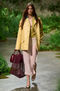 Model walking on a garden path in a pale yellow coat over a green top and pink skirt, carrying a burgundy mesh tote and matching bag.