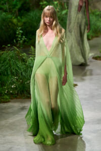 Model wearing a light green, pleated sheer gown with a deep V neckline walking an outdoor garden runway.