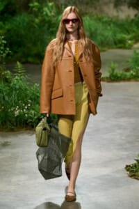Model walking outdoors in a tan leather coat, yellow top, and olive pants, carrying a green handbag and a mesh tote while wearing red sunglasses and layered necklaces.