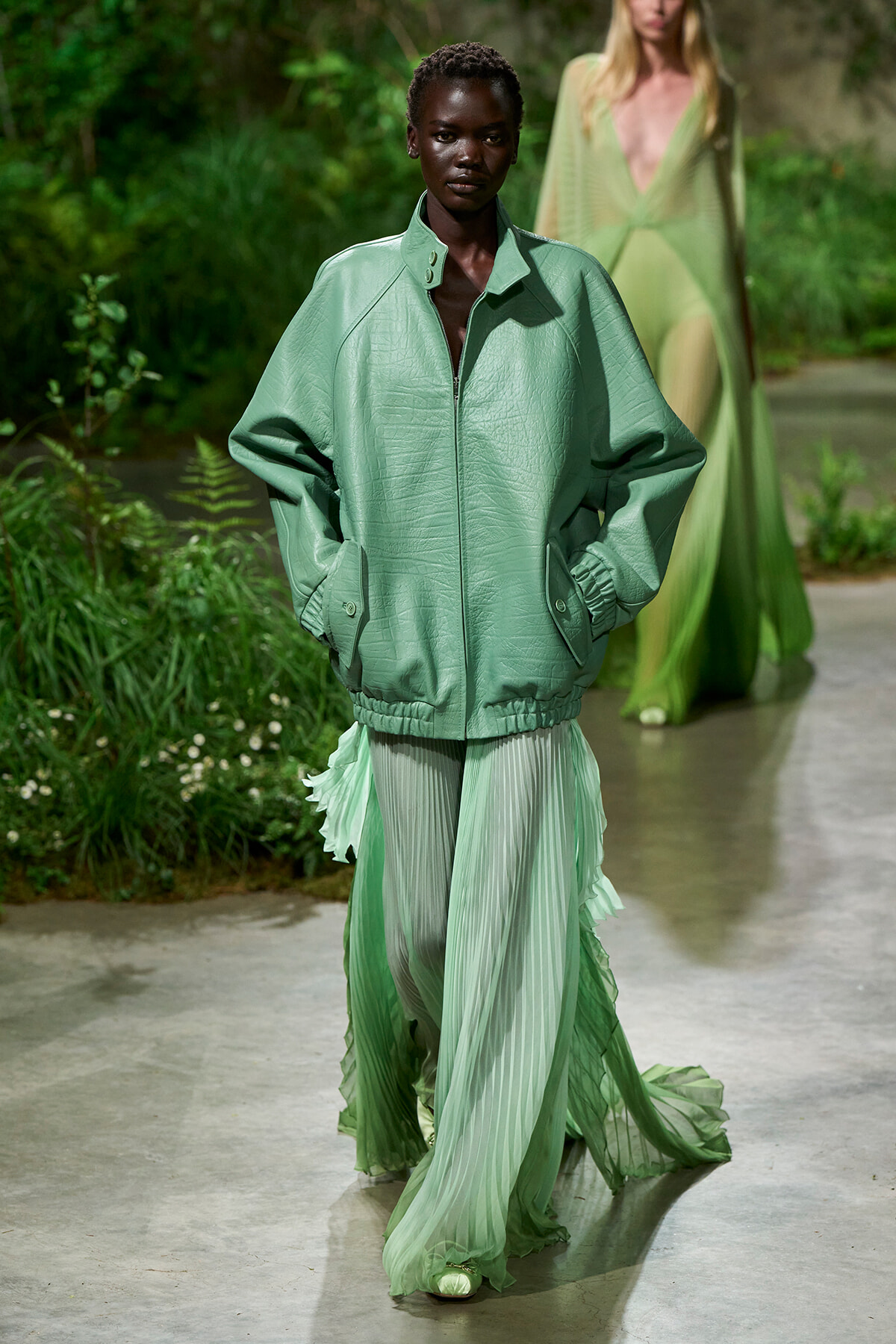 Model walks the runway in a loose mint-green leather bomber jacket over a pleated mint gown; garden backdrop on the catwalk.