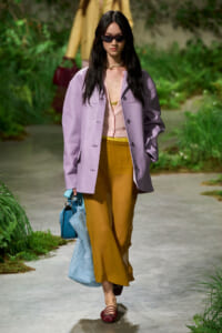 Model walking a garden runway in a pastel-lavender oversized jacket, pink blouse, mustard wide-leg pants, burgundy flats, and a blue handbag with sunglasses.