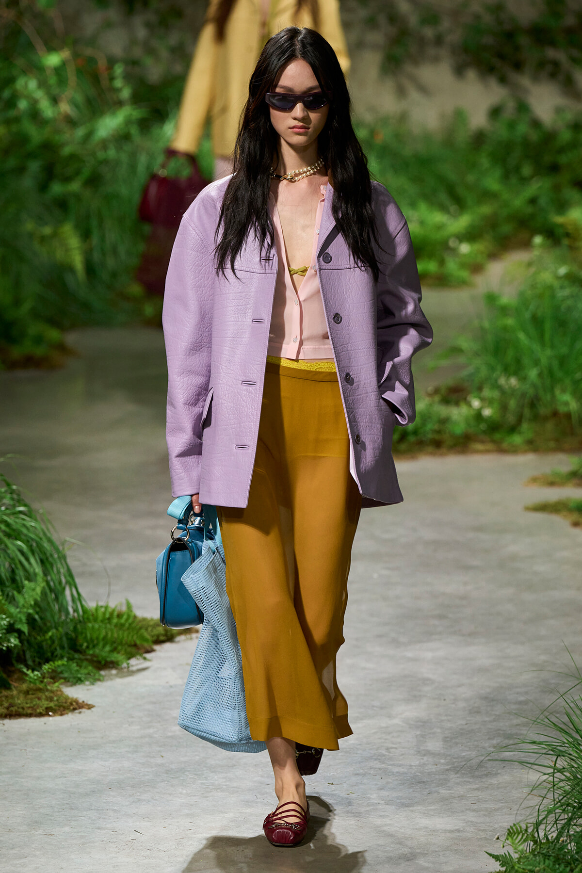 Model walking a garden runway in a pastel-lavender oversized jacket, pink blouse, mustard wide-leg pants, burgundy flats, and a blue handbag with sunglasses.