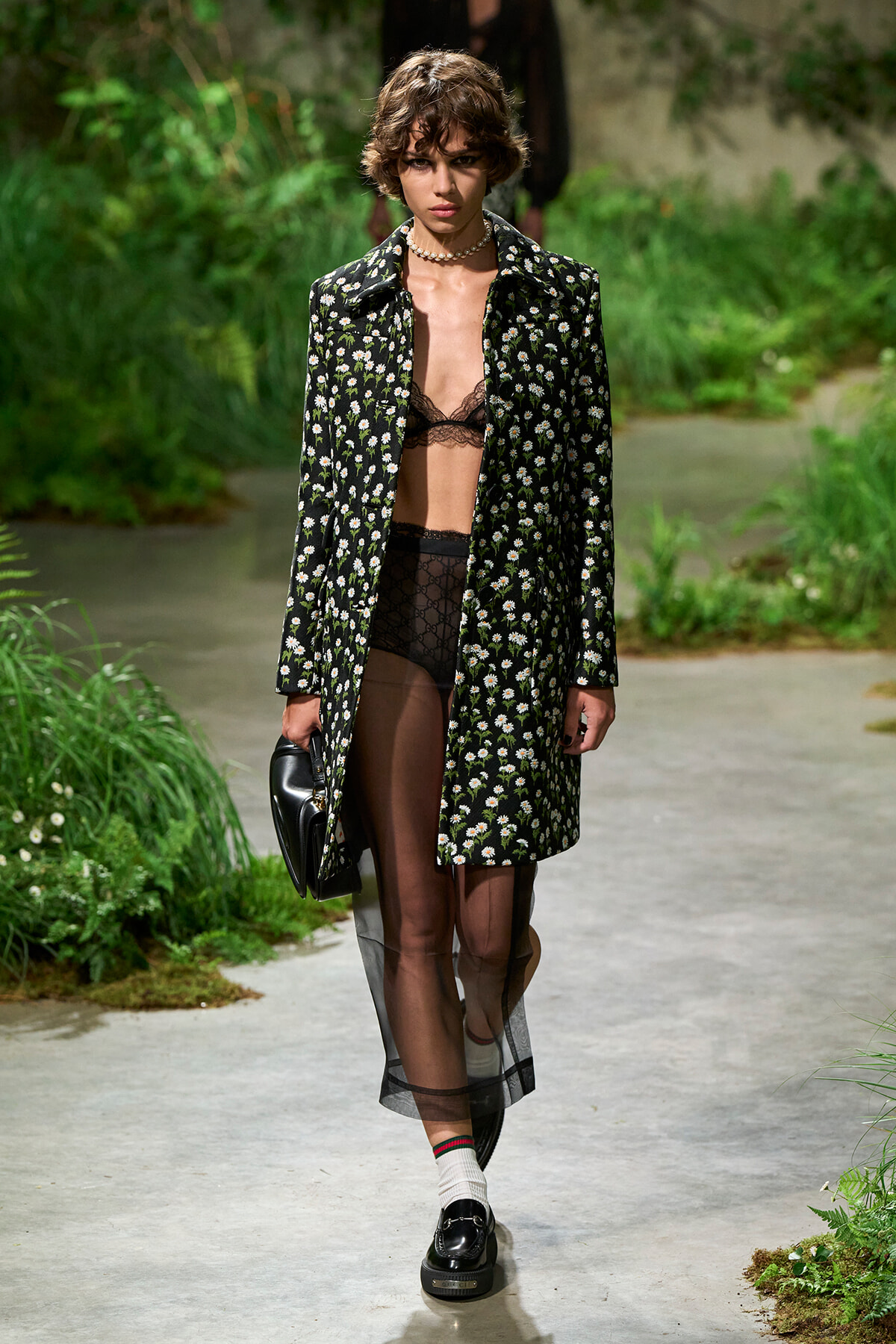 Model on a runway wearing a black floral coat over black lace lingerie, carrying a black handbag, outdoors in a garden setting.