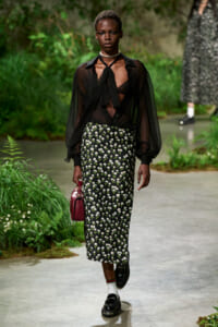 Model walking a garden-themed runway in a sheer black blouse, floral pencil skirt, burgundy handbag, and loafers with white socks.