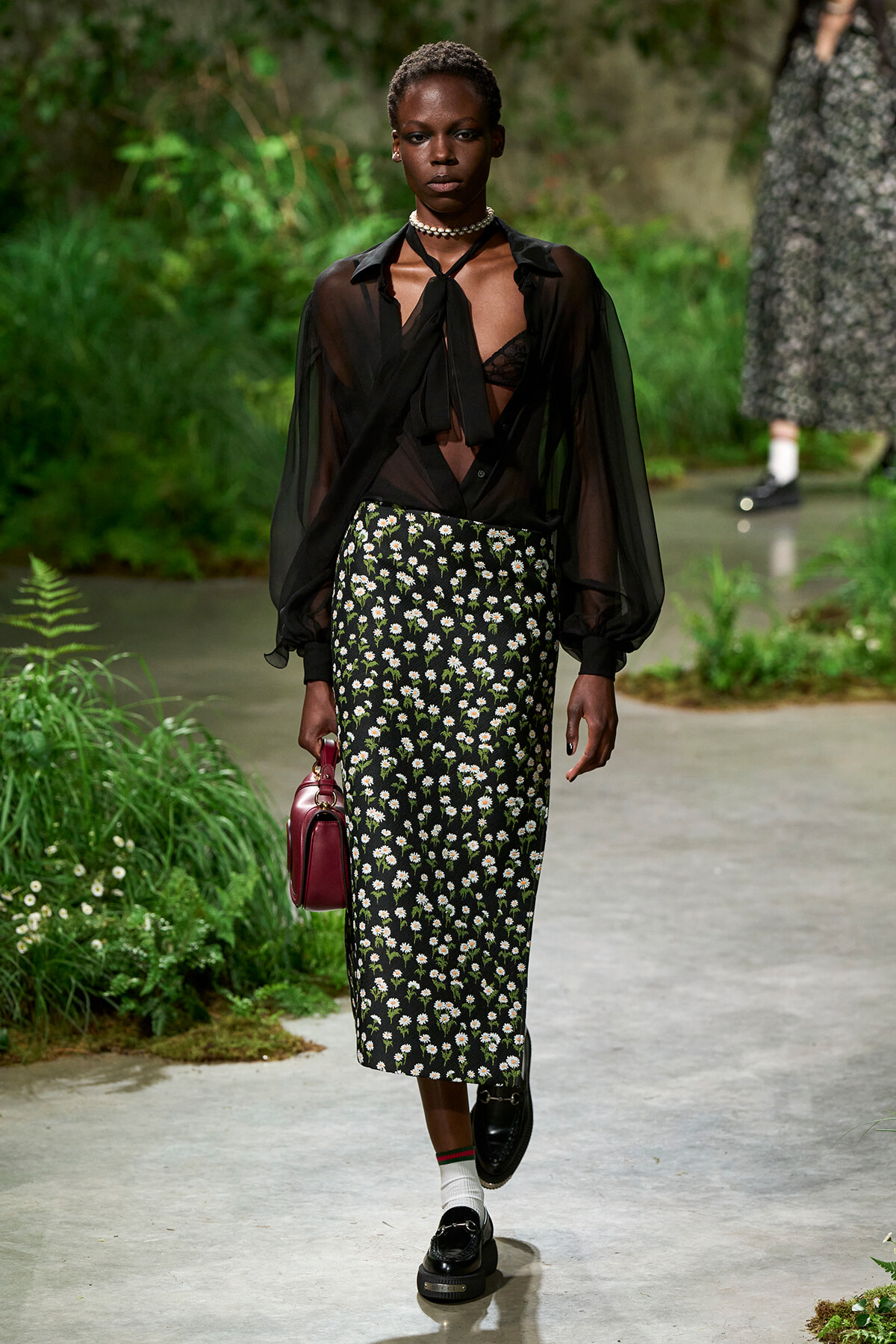 Model walking a garden-themed runway in a sheer black blouse, floral pencil skirt, burgundy handbag, and loafers with white socks.