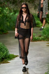 Model on a runway wearing a sheer black lingerie-style outfit with sunglasses, a pearl necklace, and a small black bag.