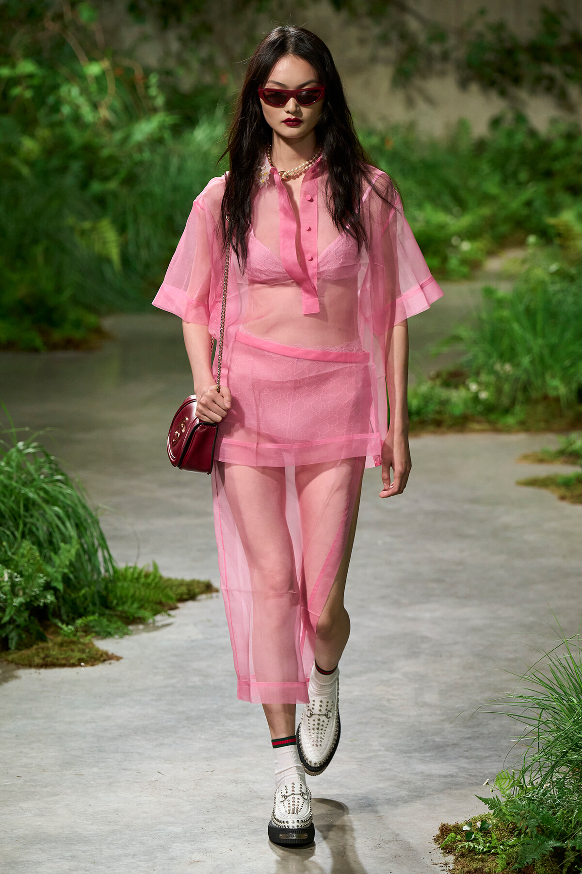 Model in pink sheer outfit with sunglasses walking along a garden path, carrying a red shoulder bag.