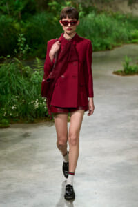Model walking along a garden path in a burgundy blazer with bow, matching shorts, sunglasses, and a shoulder bag.