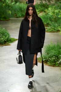 Model walks a garden runway in an oversized black coat, lace bralette, black midi skirt with a high slit, white socks, black loafers, and a small black-and-white handbag.