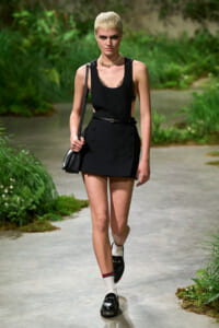 Model with short blond hair walks along a garden path in a black sleeveless top, mini skirt, white socks, black loafers, and a small black bag.