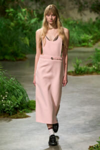 Model walking on a garden-inspired runway in a pale pink sleeveless dress with a matching belt and lace-trimmed top, wearing white socks and black loafers.