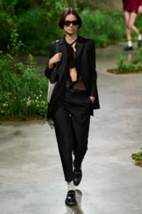 Model walking runway in a black pantsuit with sheer brown top, black blazer, sunglasses, and loafers, carrying a light-colored shoulder bag.