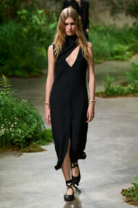 Model walking a garden runway in a black sleeveless dress with a plunging cutout and crisscross strappy heels